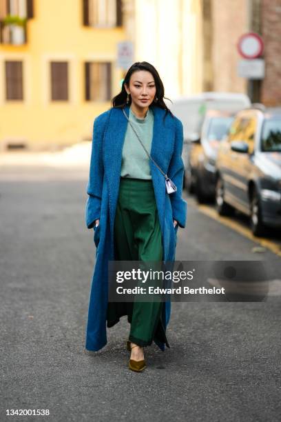 Jessica Wang wears a silver and gold pendant earrings, a pale green t-shirt, a dark green midi skirt, a blue fluffy oversized long coat, a silver...