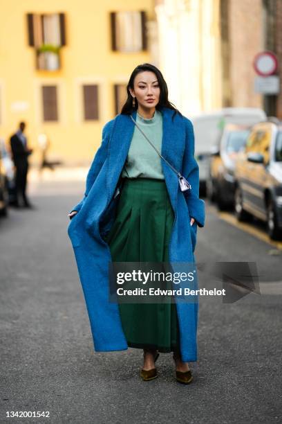 Jessica Wang wears a silver and gold pendant earrings, a pale green t-shirt, a dark green midi skirt, a blue fluffy oversized long coat, a silver...