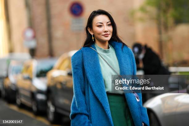 Jessica Wang wears a silver and gold pendant earrings, a pale green t-shirt, a dark green midi skirt, a blue fluffy oversized long coat, a silver...