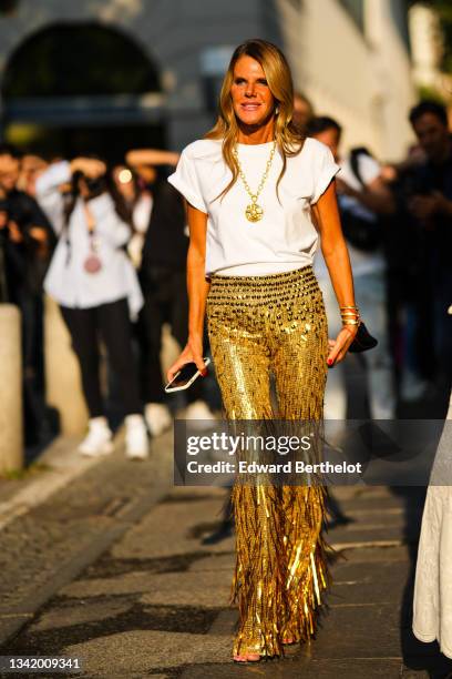 Anna Dello Russo wears a gold and white rhinestones large earrings, a gold large chain with a large pendant necklace, a white t-shirt, gold shiny...