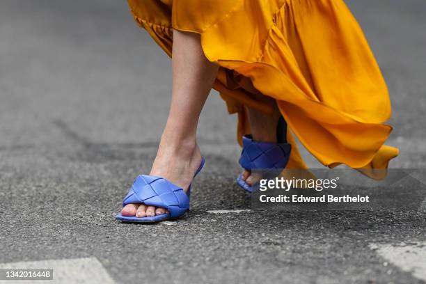 Guest wears large a yellow mustard shiny long pleated flowing asymmetric dress, blue purple quilted shiny leather block heels sandals, outside the...