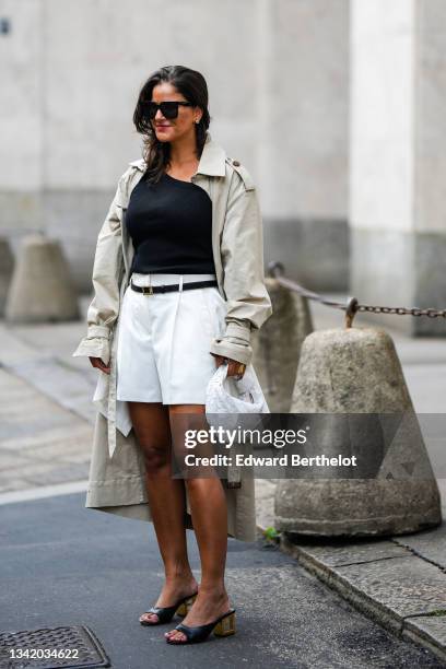 Gili Biegun wears black sunglasses, silver earrings, a black ribbed asymmetric neck t-shirt, a pale gray long oversized trench coat, a black shiny...