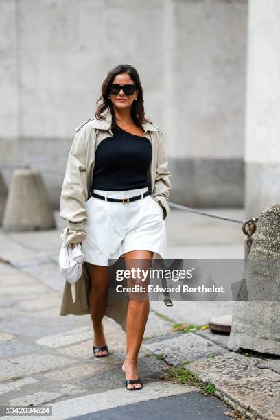 Gili Biegun wears black sunglasses, silver earrings, a black ribbed asymmetric neck t-shirt, a pale gray long oversized trench coat, a black shiny...