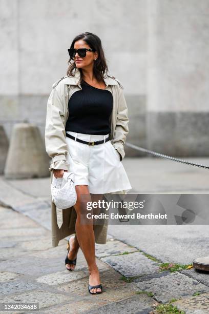 Gili Biegun wears black sunglasses, silver earrings, a black ribbed asymmetric neck t-shirt, a pale gray long oversized trench coat, a black shiny...