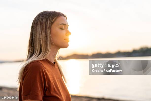 young woman meditating on the riverside, enjoying the last sun rays of the day - breathing exercise stockfoto's en -beelden