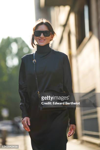Guest wears black sunglasses, silver earrings, a black silk turtleneck / long sleeves flowing long dress, a black shiny leather Jacquemus crossbody...