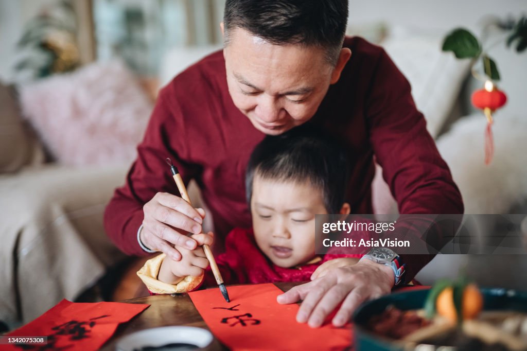 Grandfather practising Chinese calligraphy for Chinese New Year Fai Chun (Auspicious Messages) and teaching his grandson by writing on couplets at home