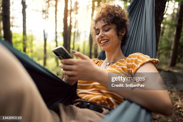 cheerful female hiker resting in a hammock in the pine forest and using a smart phone - hammock phone stock pictures, royalty-free photos & images