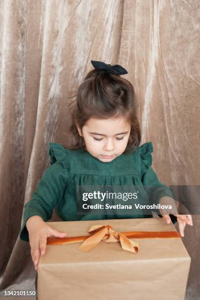 a toddler girl in a green linen dress and a headband  holds a christmas present wrapped in kraft paper and decorated with a velour ribbon and looks at it within temptation. christmas outfit. preparing for celebration. festive season. lifestyle portrait. - toddler girl dress stockfoto's en -beelden