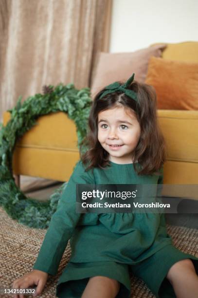 smiling little girl in green linen clothes and a headband sits on the floor near the yellow fabric sofa and christmas wreath at home. christmas mood. getting ready for the holiday. festive season. - merry christmas in armenian 個照片及圖片檔