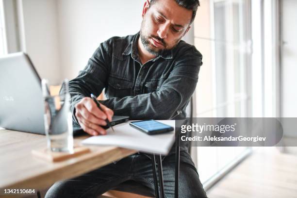 focused serious male working from home on laptop and smartphone while writing down data - journalist computer stock pictures, royalty-free photos & images