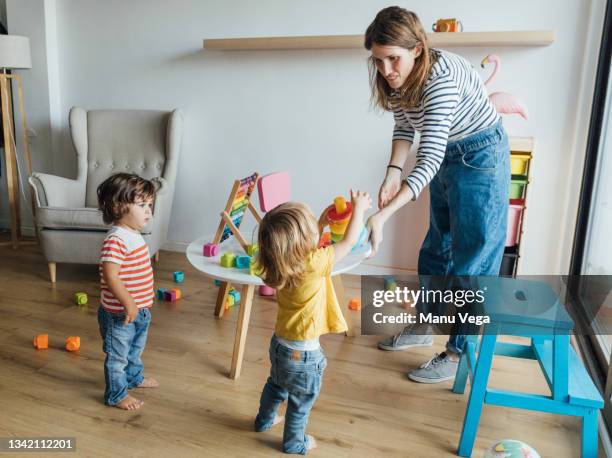 positive young female babysitter in casual clothes playing with infant children in cozy playroom with colorful educational toys - nursery school stockfoto's en -beelden