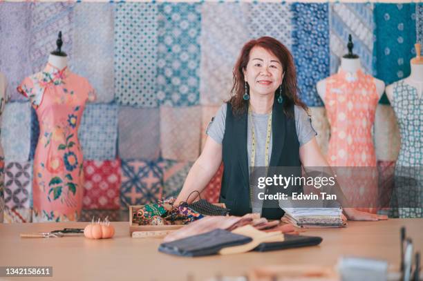 malaysian chinese mature woman boutique small business owner portrait in front of her batik cheongsam fabric textile collection retail store looking at camera smiling - malaysia batik stock pictures, royalty-free photos & images