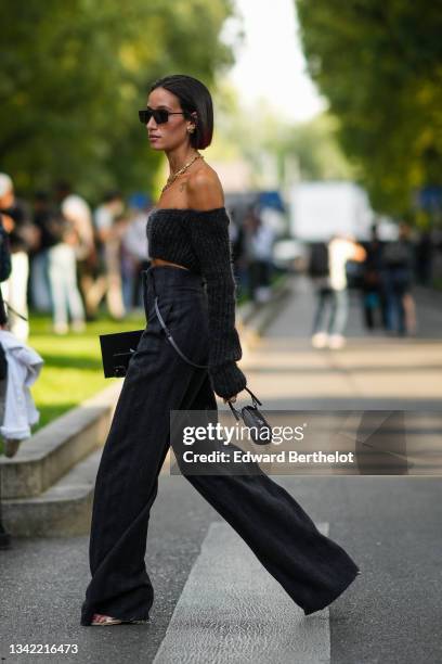 Alexandra Guerain wears black sunglasses, earrings, gold chain necklaces, a dark gray ribbed off-shoulder / long sleeves / cropped pullover, a dark...