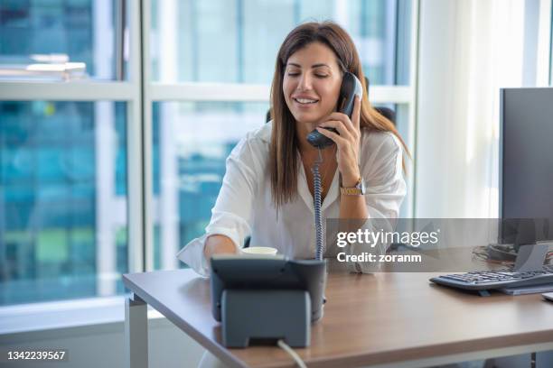 businesswoman using landline at work and smiling at her desk - vaste telefoon stockfoto's en -beelden