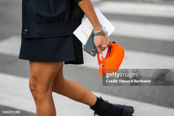 Guest wears a black coat, a black dress, a neon orange leather small Coperni handbag, a silver watch, a gold ring, black shiny leather Prada ankle...