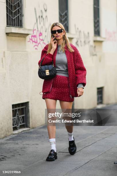 Natalia Verza wears black sunglasses, a pale gray polo shirt, a red shiny braided leather zipper jacket, a matching red shiny braided leather short...