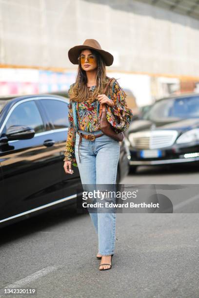 Guest wears gold earrings, brown YSL sunglasses from Yves Saint Laurent, a brown felt / wool hat, a multicolored print patter ruffled shirt, a brown...