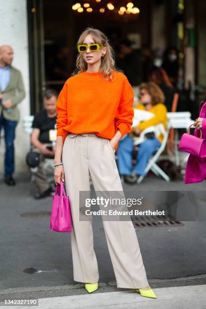 Guest wears yellow sunglasses, a neon orange wool pullover, beige high waist suit pants, gold and brown earrings, pearls bracelets, a neon shiny...
