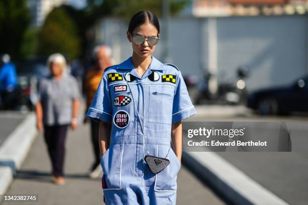 Guest wears sunglasses, silver earrings, a baby blue patched by black / white / yellow pattern cargo jumpsuit from Miu Miu, a black shiny leather...