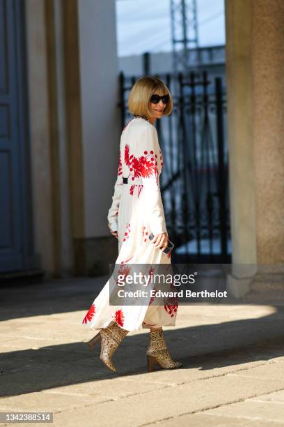 Anna Wintour wears black sunglasses from Chanel, a gold and silver chain necklace, a white with flower print pattern long sleeves / belted / flowing...