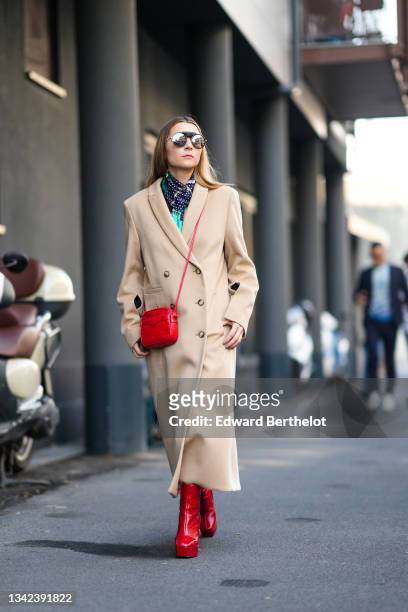 Julia Comil wears sunglasses, a green / blue with white polka dots silk scarf, a beige wool long coat, a red shiny leather crocodile pattern...