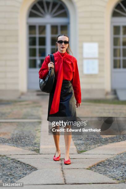 Guest wears black sunglasses, a red oversized wrap shirt with a knotted neck, a black shiny leather shoulder bag, a black shiny leather high waist...