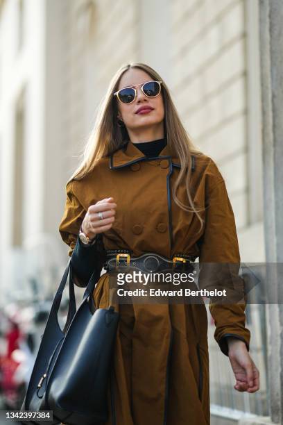 Xenia Tchoumitcheva wears black sunglasses, a black turtleneck pullover, a camel buttoned long oversized slit / split coat, a black shiny leather...