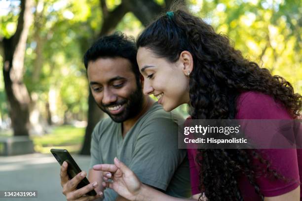 happy diverse friends at the park sitting on a bench close together looking at social media on smartphone talking and smiling - happy couple using cellphone stockfoto's en -beelden