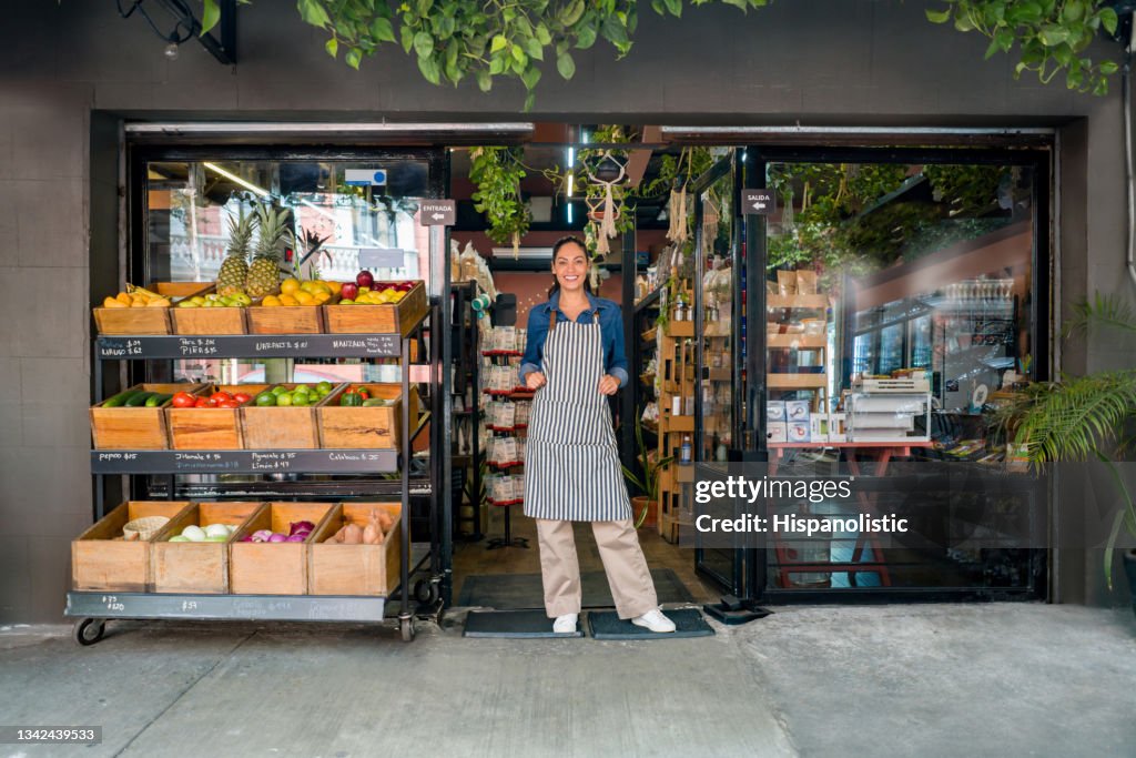 Happy woman working at a local food market and standing at the door waiting for clients