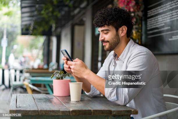 hombre feliz bebiendo revisando su teléfono celular en una cafetería mientras bebe un capuchino - cafe outside fotografías e imágenes de stock