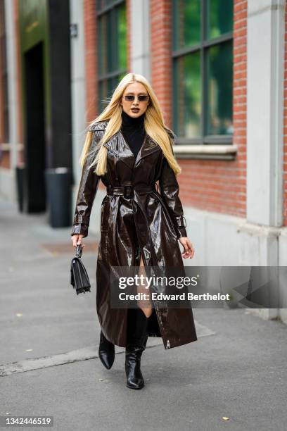 Guest wears sunglasses, a black wool turtleneck dress, a brown shiny leather / vinyl long belted trench coat, a black shiny leather handbag, a black...