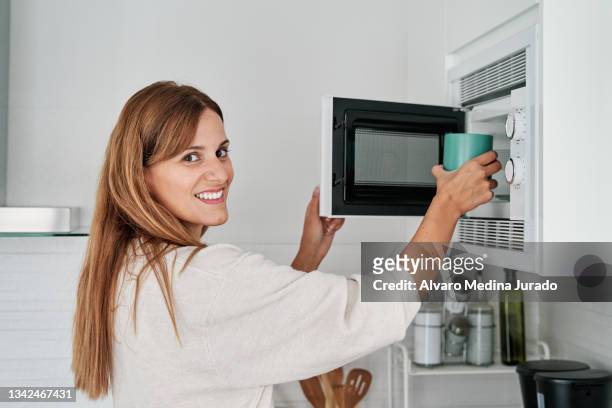 pretty young woman in the kitchen putting a cup in the microwave to heat it up. - microwave dish stock pictures, royalty-free photos & images