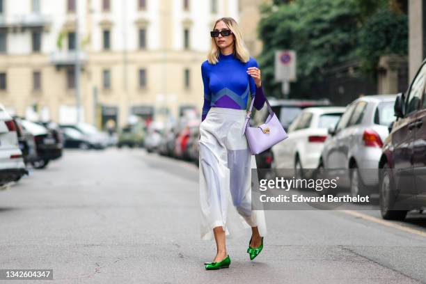 Candela Novembre wears black sunglasses, a royal blue electric with purple and gray print pattern ribbed long sleeves high neck jumpsuit, a white...