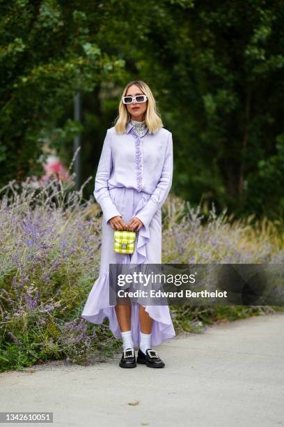 Candela Novembre wears white sunglasses, a silver sequined turtleneck t-shirt, a pale purple buttoned shirt, a pale purple flowing ruffled asymmetric...