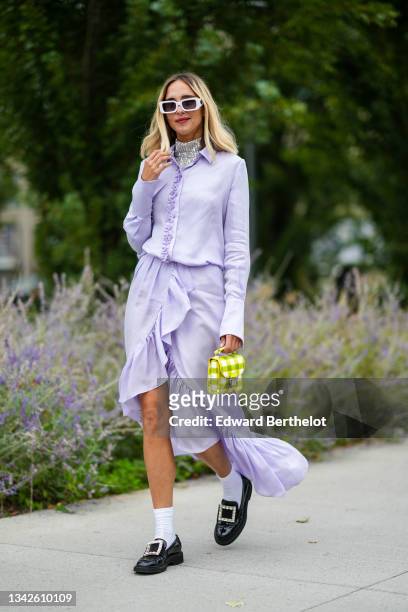 Candela Novembre wears white sunglasses, a silver sequined turtleneck t-shirt, a pale purple buttoned shirt, a pale purple flowing ruffled asymmetric...