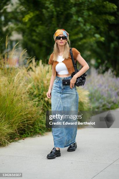 Natalia Verza wears a beige with red / blue / orange print pattern scarf, black sunglasses, a white cropped t-shirt, a brown fluffy open t-shirt...