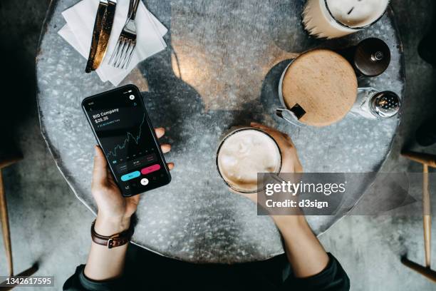 overhead view of young woman enjoying a coffee break, managing finance and investment, checking financial stock market data on smartphone in cafe. smart banking with technology - rise above it stock pictures, royalty-free photos & images