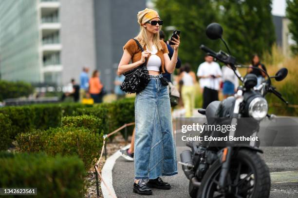 Natalia Verza wears a beige with red / blue / orange print pattern scarf, black sunglasses, a white cropped t-shirt, a brown fluffy open t-shirt...