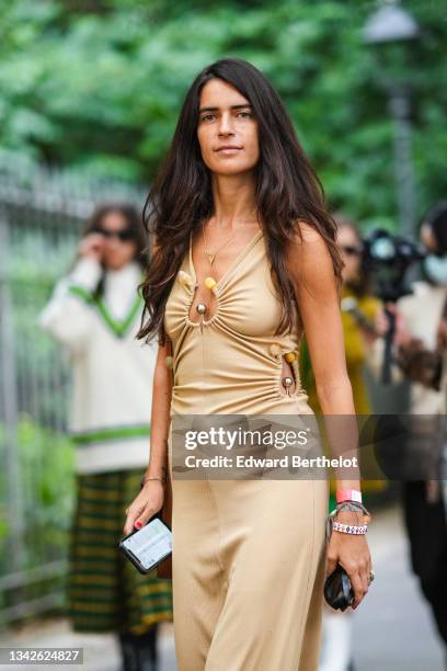 Chiara Totire wears a gold chain pendant necklace, a beige ribbed V-neck / cut-out flowing long dress with silver and yellow stones, a white with...