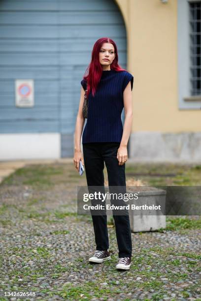 Model wears a dark blue and dark green ribbed cool sleeveless pullover, black flared denim jeans pants, a brown with white C logo in coated canvas...