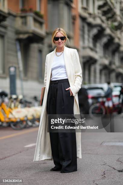 Lisa Aiken wears black Ray Ban sunglasses, silver earrings, a gold chain necklace, a white ribbed t-shirt, a white latte long coat, black palazzo...