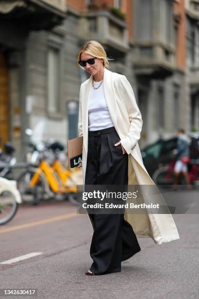 Lisa Aiken wears black Ray Ban sunglasses, silver earrings, a gold chain necklace, a white ribbed t-shirt, a white latte long coat, black palazzo...