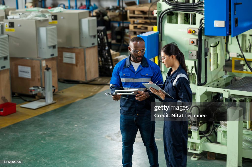 Industrial and Manufacturing Engineering, Quality Control and Management in a Production Process. African supervisor explaining to Asian junior engineer by engineering parts. They are discussing in front of mechanical press machine over component parts.