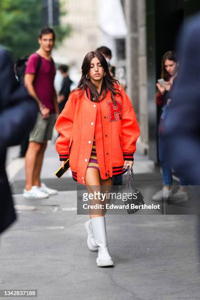 Guest wears gold earrings, a burgundy with yellow stripes short dress, a neon orange sport oversized bomber coat from Philosophy, a black shiny...