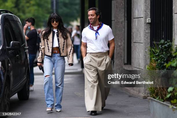 Guest wears black sunglasses, a white cropped t-shirt, a beige with black print pattern oversized cropped jacket, a brown shiny leather belt, blue...