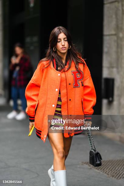 Guest wears gold earrings, a burgundy with yellow stripes short dress, a neon orange sport oversized bomber coat from Philosophy, a black shiny...