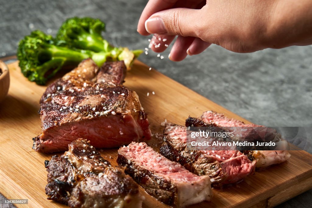 Top view of a hand placing coarse salt on a freshly cooked veal entrecote on a wooden board.