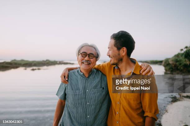 senior father and adult son having a good time on beach at sunset - asian smiling father son stock-fotos und bilder