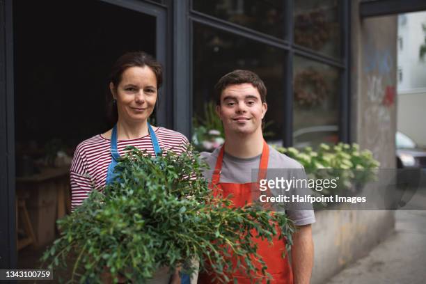 young male florist with down syndrome with colleague standing outdoors in front of flower shop, looking at camera. - dedication stock pictures, royalty-free photos & images